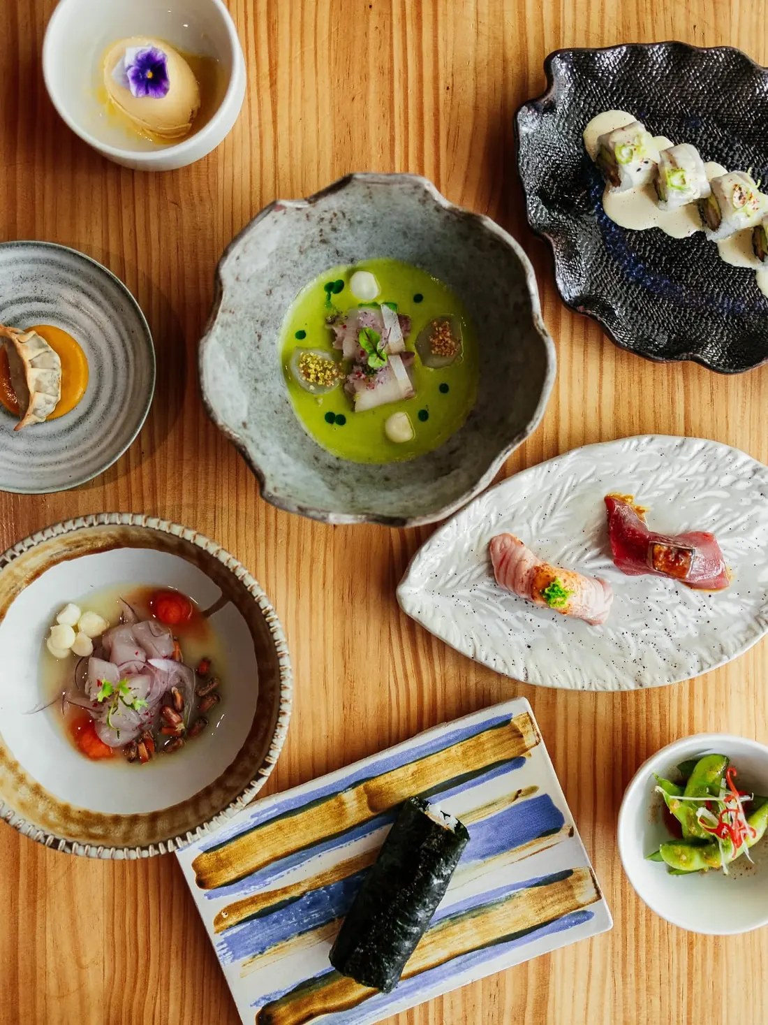 Wooden table seen from above with tiradito, nigiri, gyoza, a hand roll, ceviche and edamame