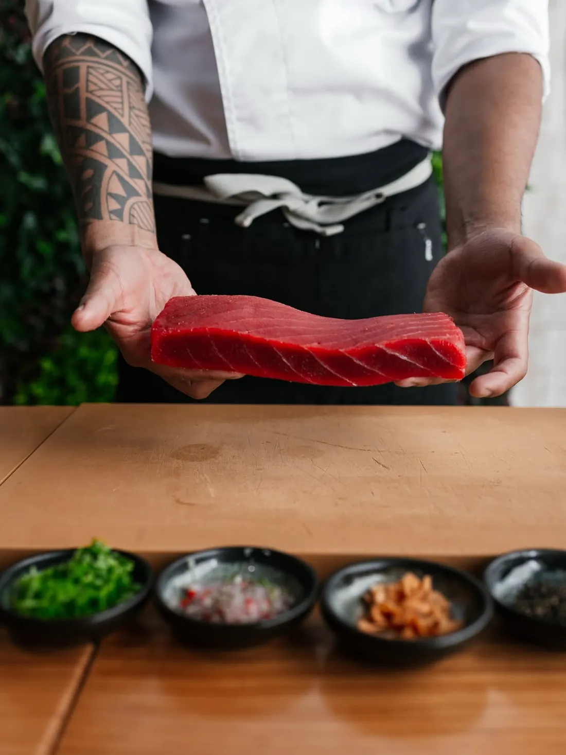 Two hands holding a block of bluefin tuna over the board, with bowls of condiments in front