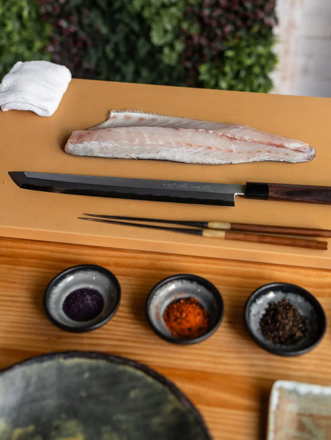 A white fish fillet on the chopping board, next to a Japanese knife, chopsticks and three bowls of spices